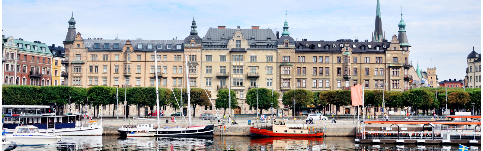 A large, beige building with many windows, about six stories tall, near a waterway lined with boats.